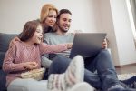 father, wife, and daughter in a living room in front of a laptop eating popcorn