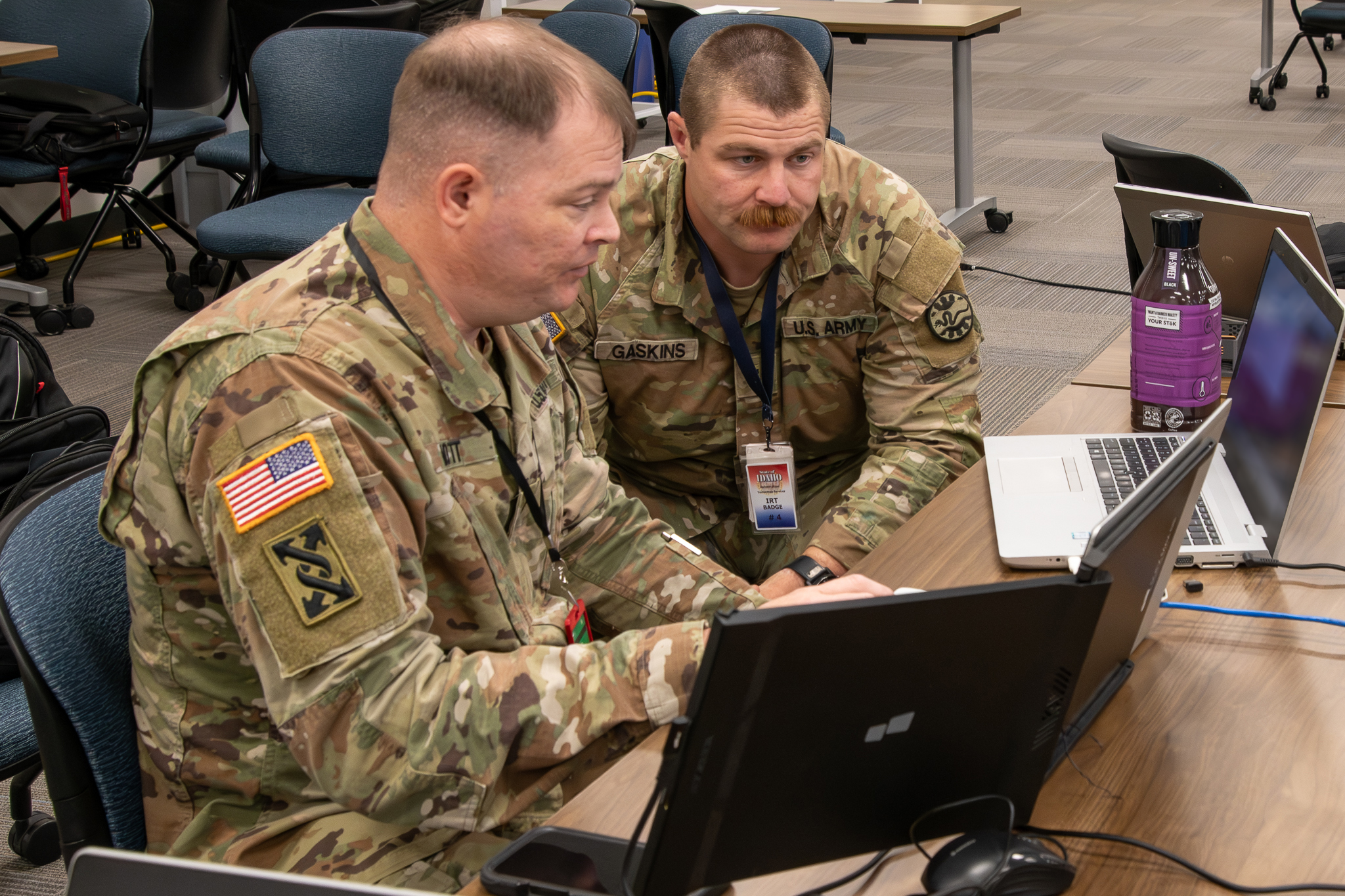 Two individuals in uniform are sitting at a table working together on laptops.
