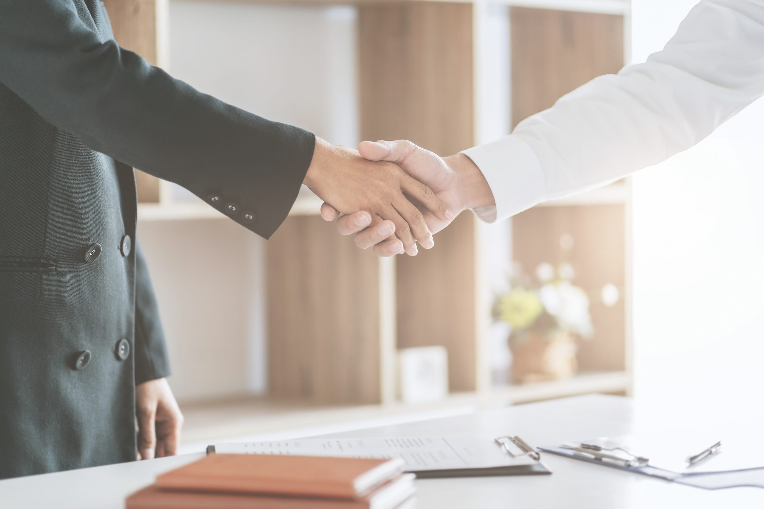 Two people in business attire shaking hands across a desk with documents and a clipboard, in a bright office setting.